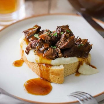 A close-up view of a slow cooker beef manhattan with brown gravy drizzled over the top of the bread, potatoes, and shredded roasted beef all on a white dinner plate.