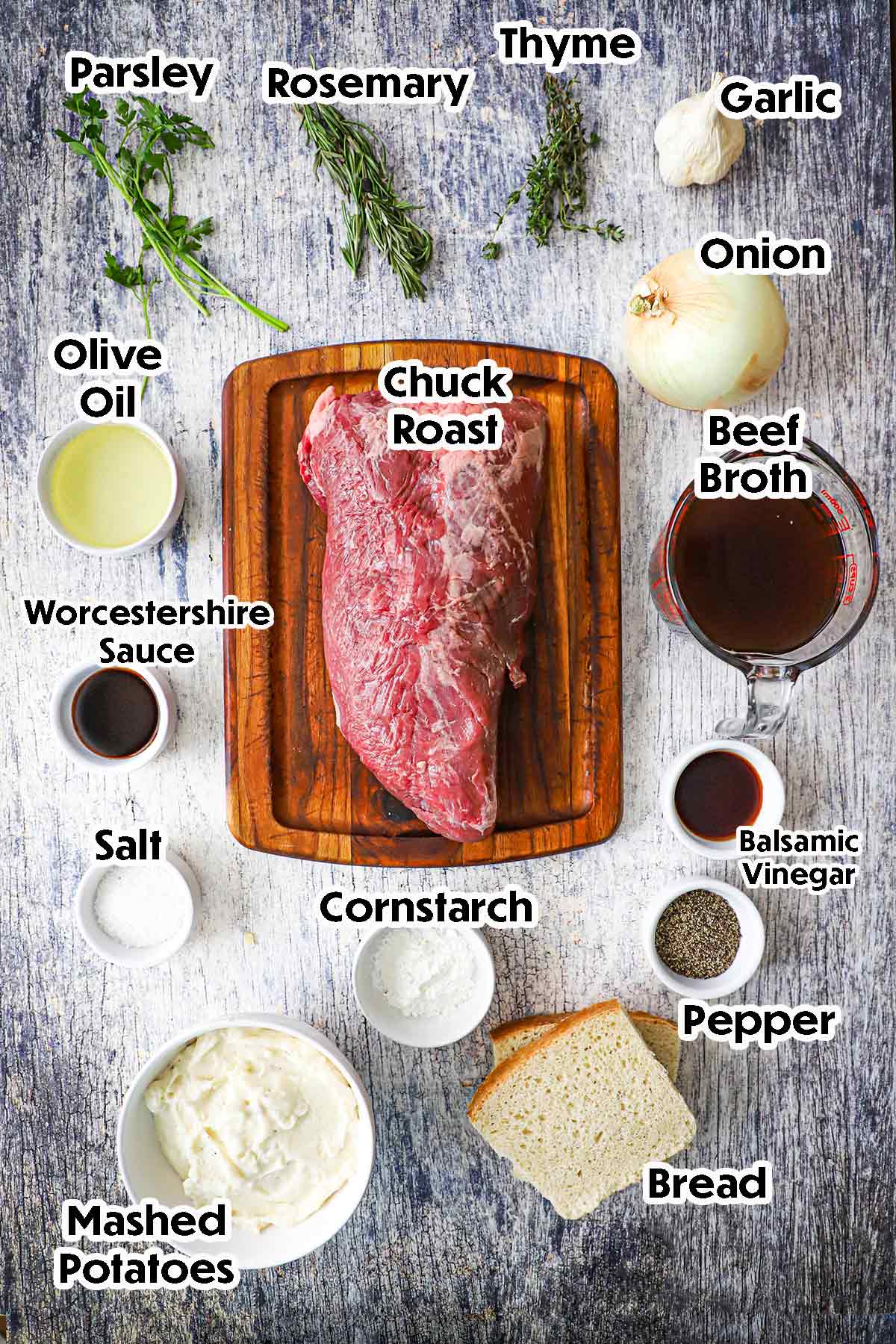 An arrangement of ingredients for slow cooker beef Manhattan on a grey wooden background including a chuck roast, beef broth, onion, garlic, fresh herbs, Worcestershire sauce, vinegar, mashed potatoes, bread, and seasonings.