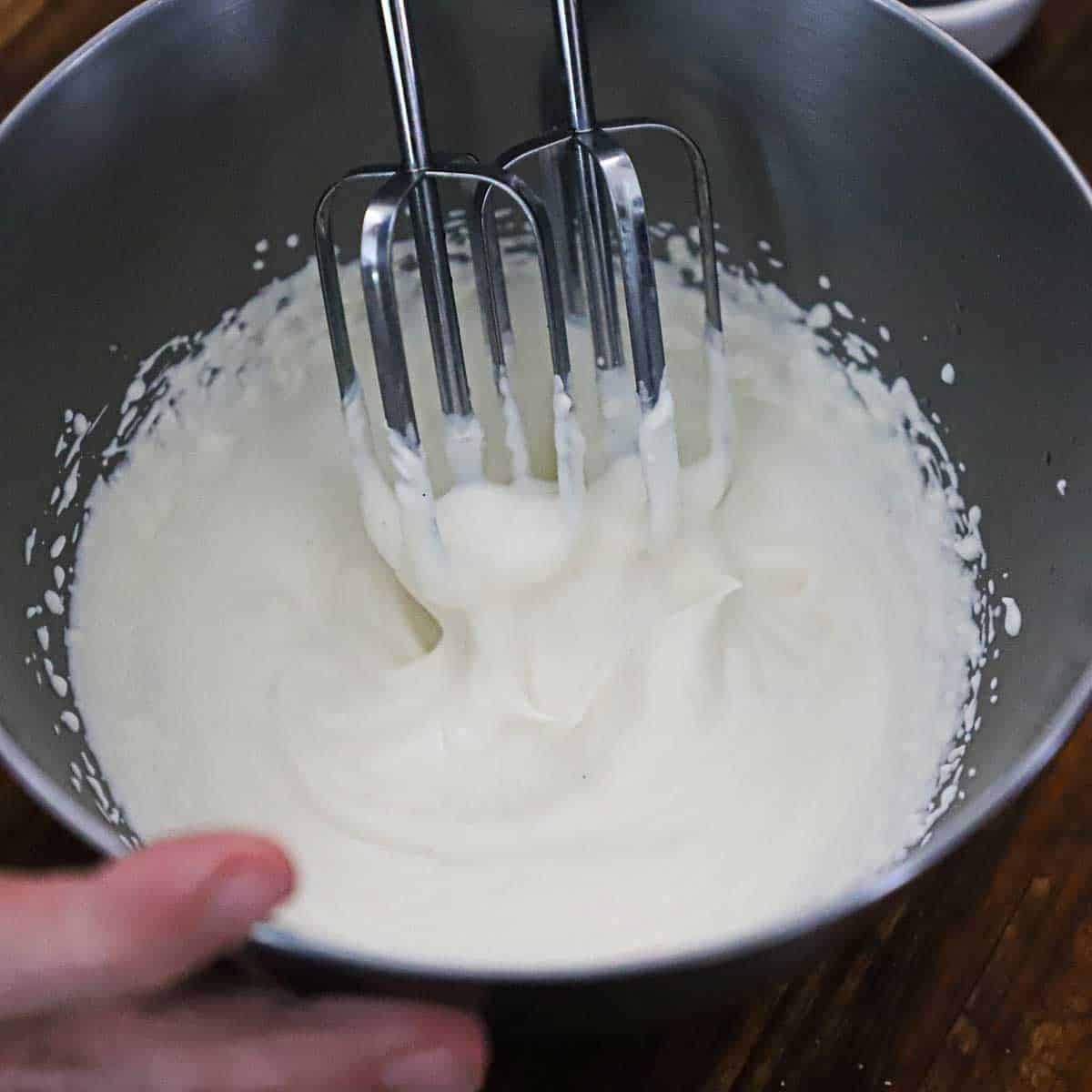 A person using an electric hand mixer to whip mascarpone cheese and heavy cream into soft peaks in a metal bowl.