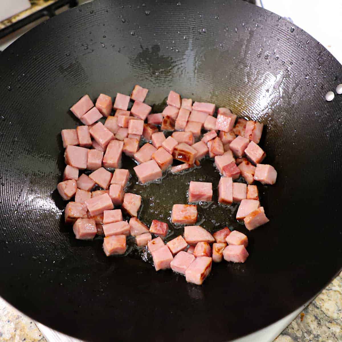 Pieces of cubed left-over ham being lightly fried in peanut oil in a large wok on a gas stove.