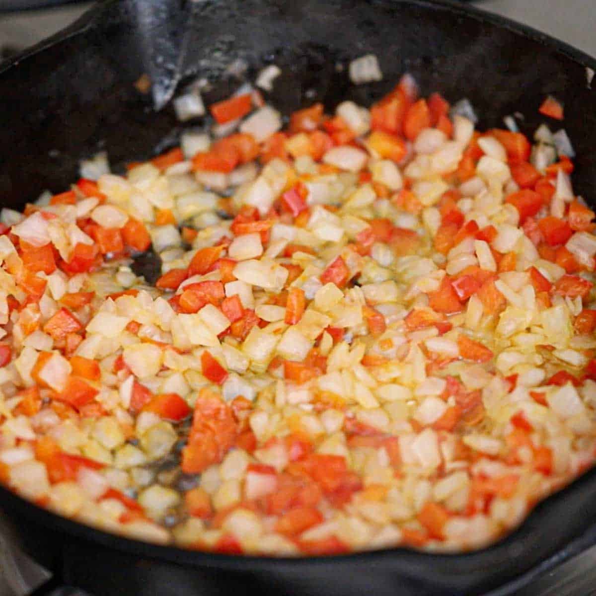 A close-up view of chopped red bell pepper and onion being sautéed in a cast-iron skillet on a gas stove.