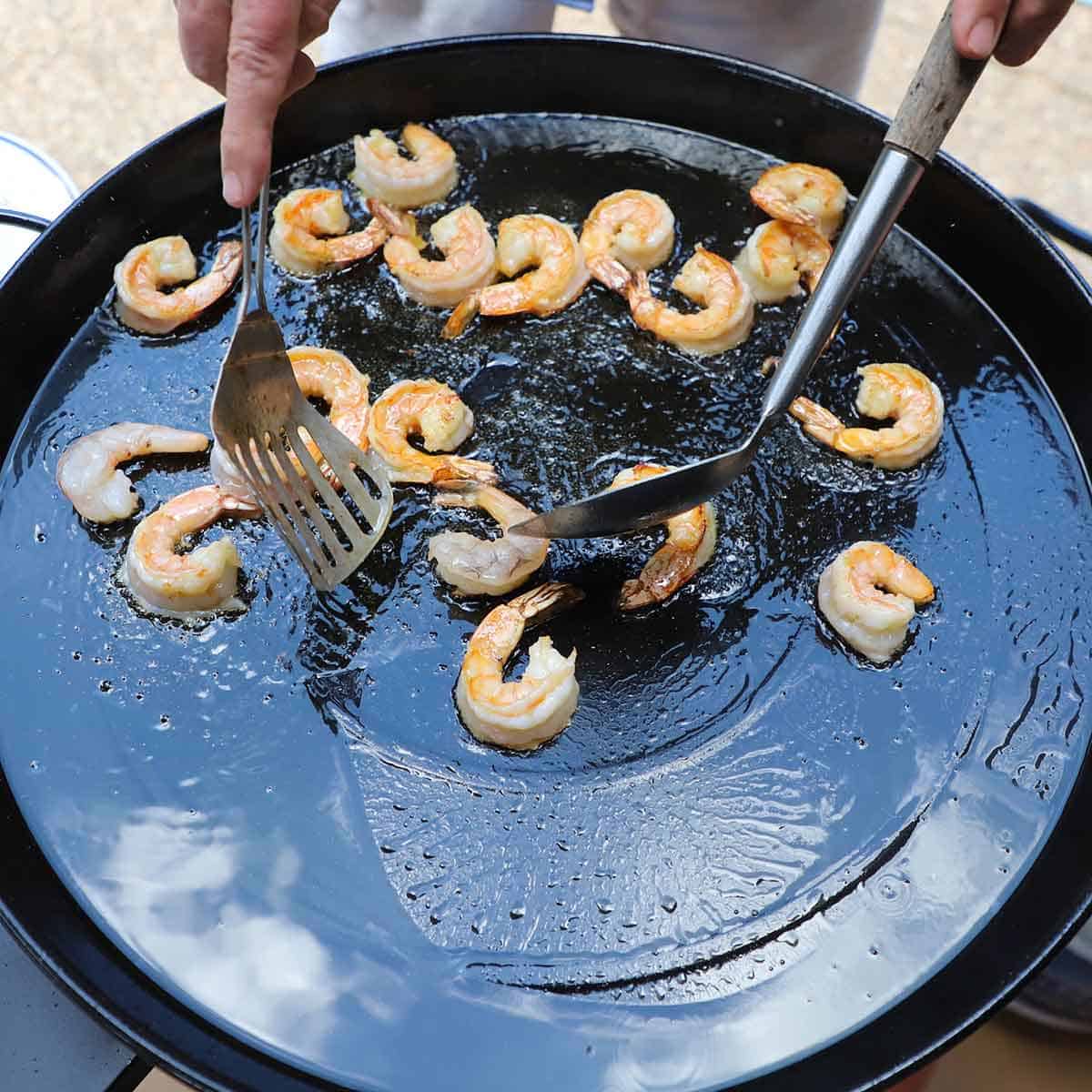 A person using two metal spoons to toss shrimp in hot oil in a large paella pan.