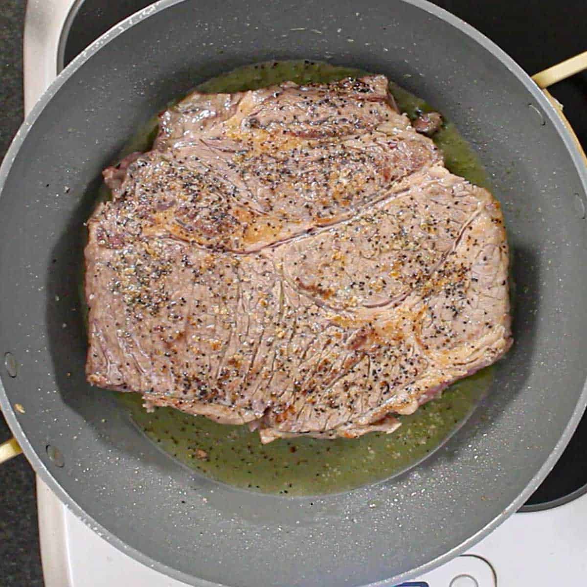 A large beef chuck roast that has been seasoned with salt and pepper and is being seared in oil in a large non-stick skillet.