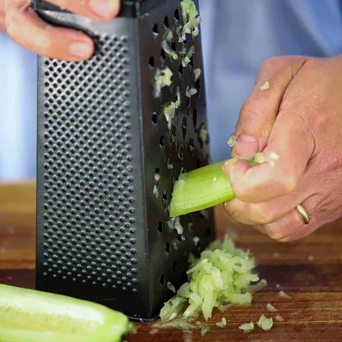 A person using a box grater to shred a cucumber that has been peeled, seeded, and sliced in half, lengthwise.