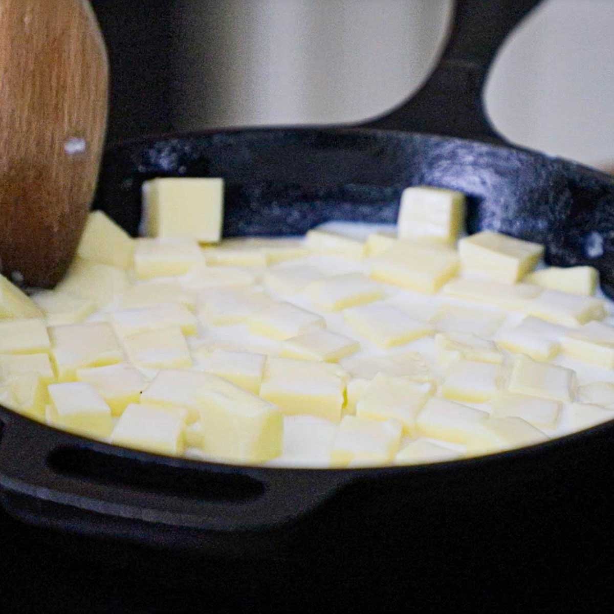 A person using a wooden spoon to stir cubed chunks of white American cheese, milk, and water over low heat in a black cast-iron skillet.