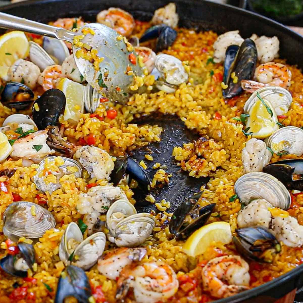 A person using a large serving spoon to serve authentic Spanish seafood paella from a large pan and revealing the socarrat on the bottom of the rice.