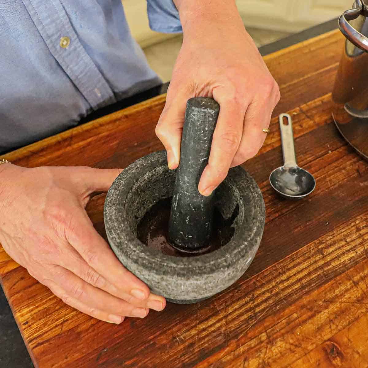 A person using a mortar and pestle to grind saffron threads with warm shellfish stock on a wooden cutting board.