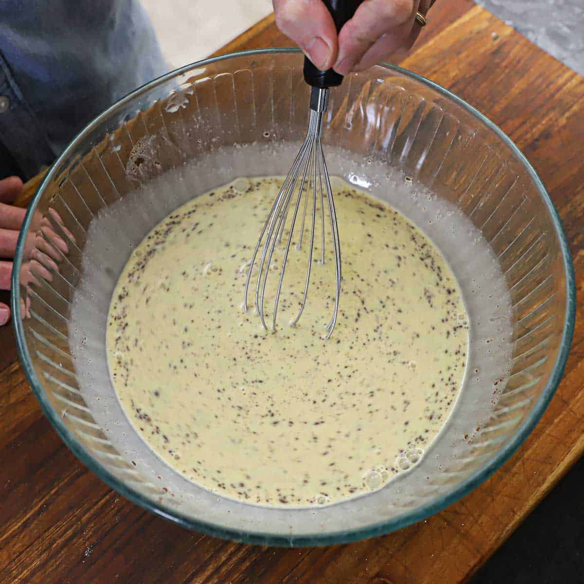 A person using a whisk to mix an egg and cinnamon custard in a glass bowl for caramelized brioche French toast.