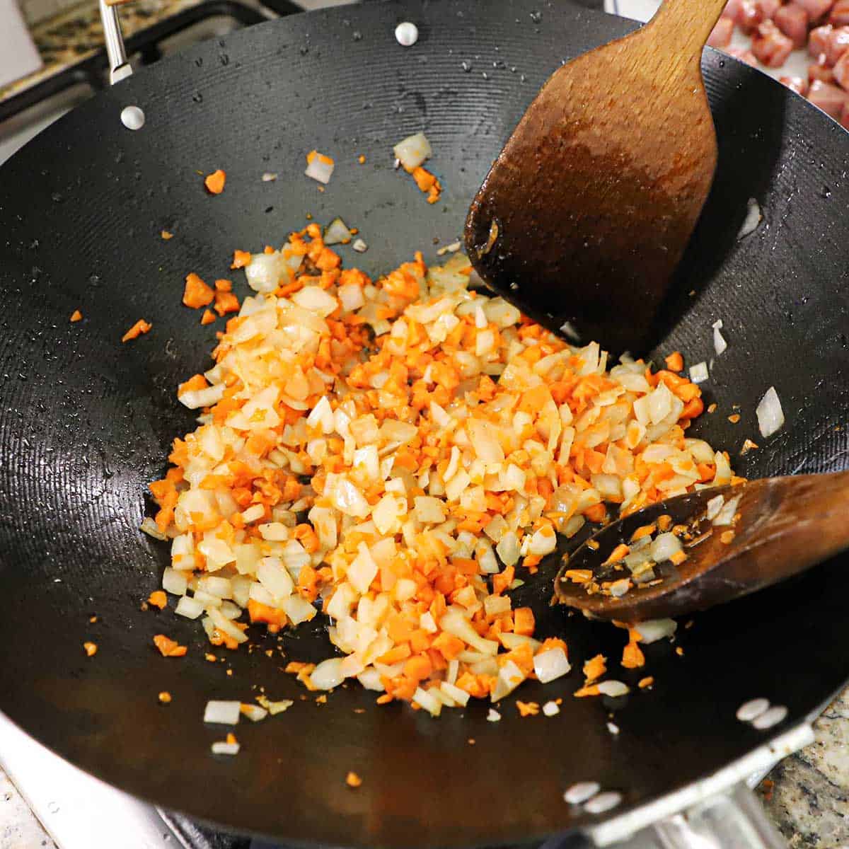 A person using two wooden spoons to stir simmering chopped onion and carrots in oil in a large wok on a gas stove.