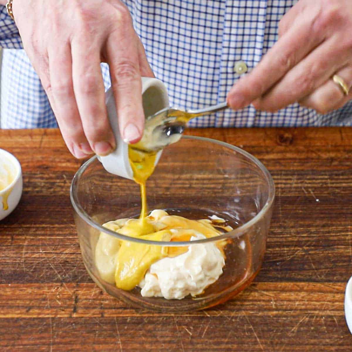 A person using a spoon to transfer yellow mustard from a small bowl into a larger glass bowl containing mayonnaise, honey, and Dijon mustard.
