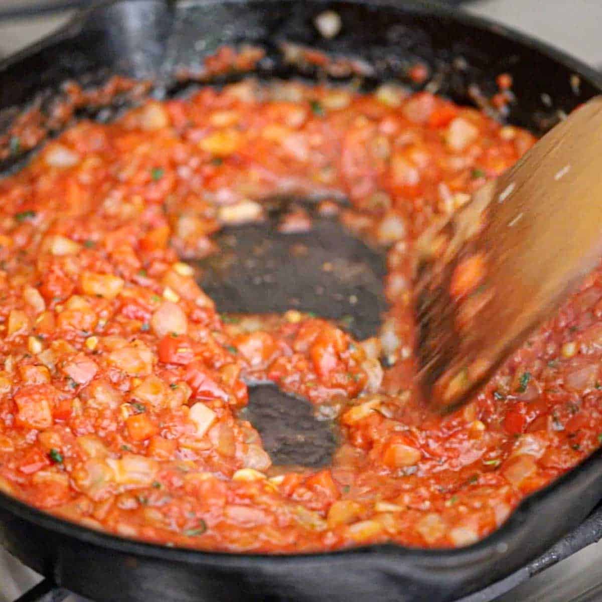 A person using a wooden spatula to stir simmering crushed tomatoes along with sautéed onions and red bell pepper in a cast-iron skillet on a gas stove.