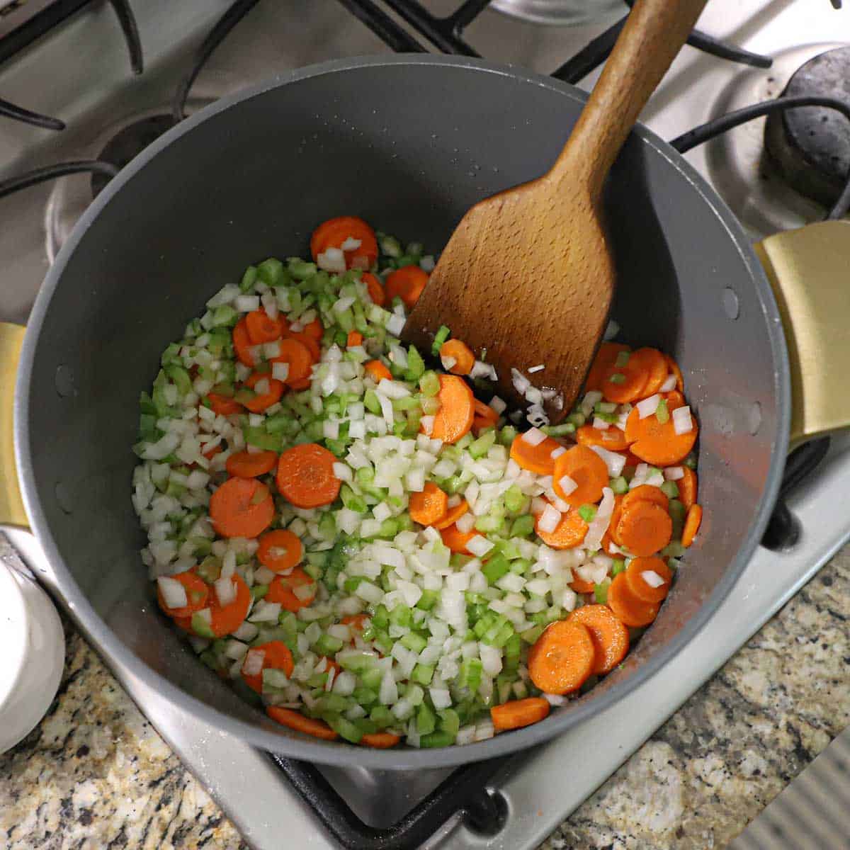 A person using a large spatula to stir chopped onions, carrots, and celery that are being sautéed in a large stock pot on a gas stove.