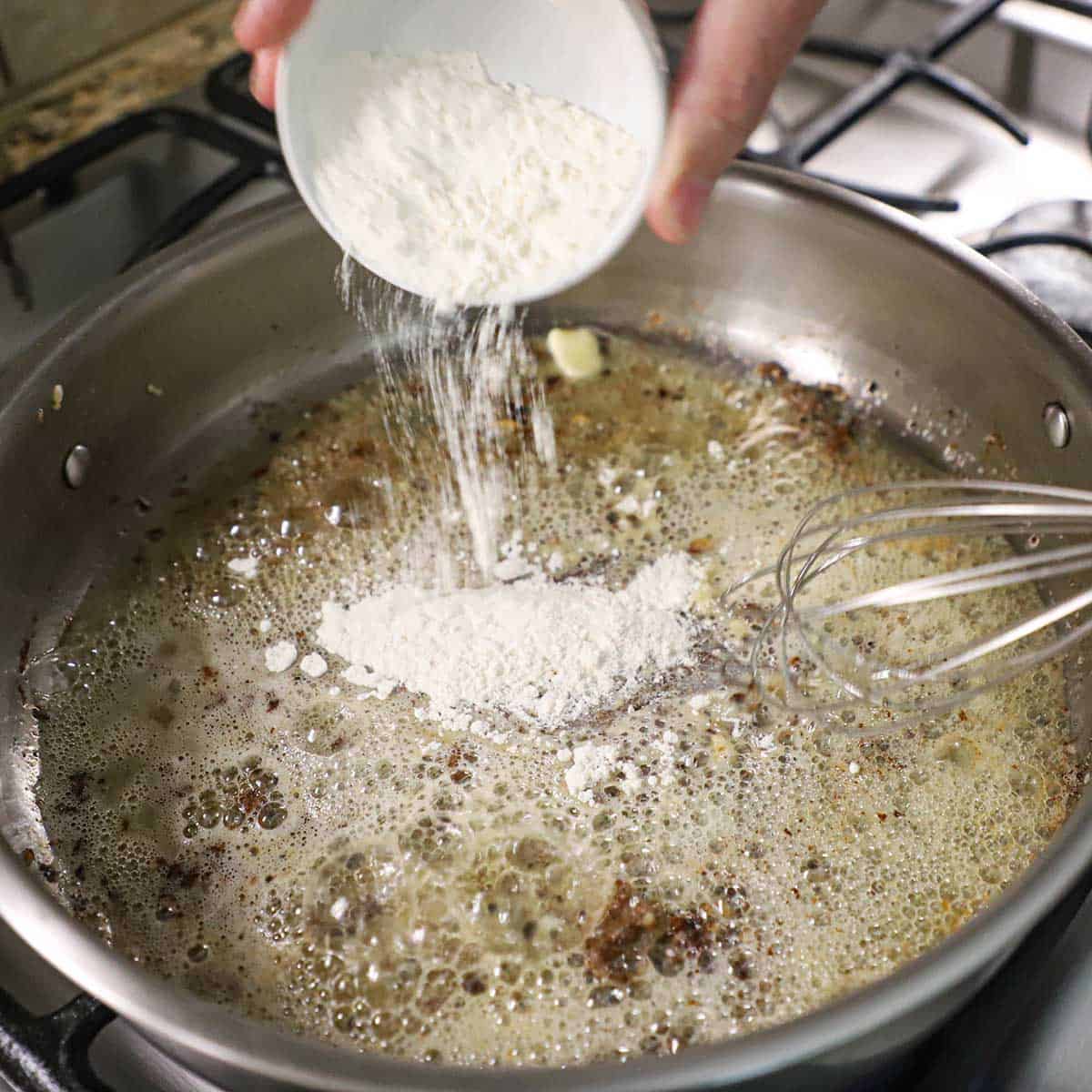 A person transferring all-purpose flour from a small white bowl into a large skillet that is filled with a simmering roux on a gas stove.