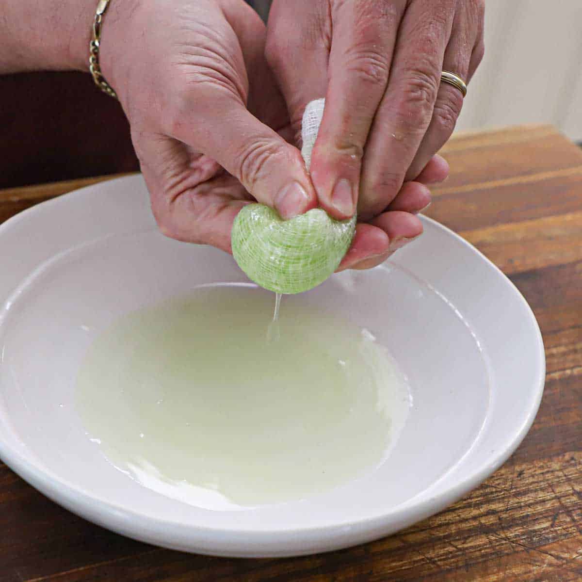 A person squeezing water from shredded cucumber that is wrapped in cheesecloth over a shallow white bowl.