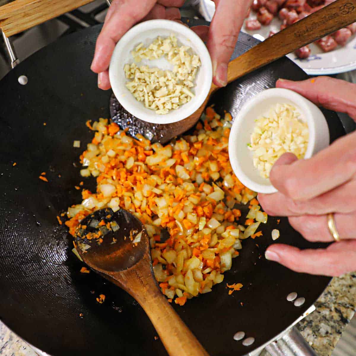 A person holding a small bowl of minced ginger in one hand and a small bowl of minced garlic over a wok of sautéed chopped onion and carrots.