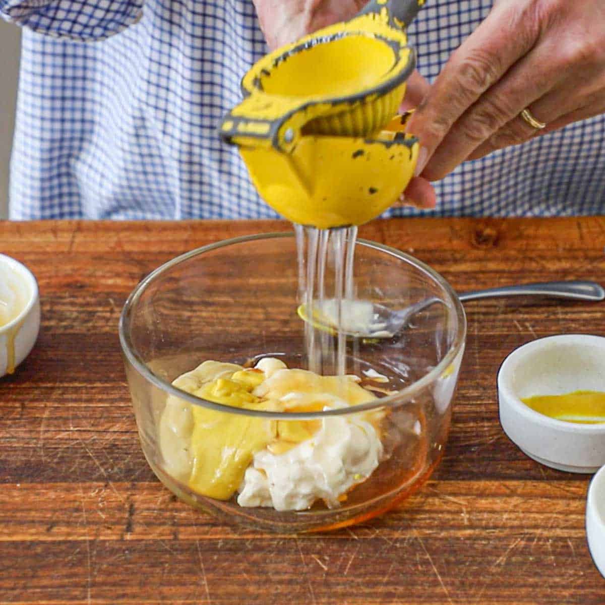 A person using a citrus juicer to squeeze fresh lemon juice into a glass bowl filled with mayonnaise, honey, Dijon mustard, and yellow mustard.