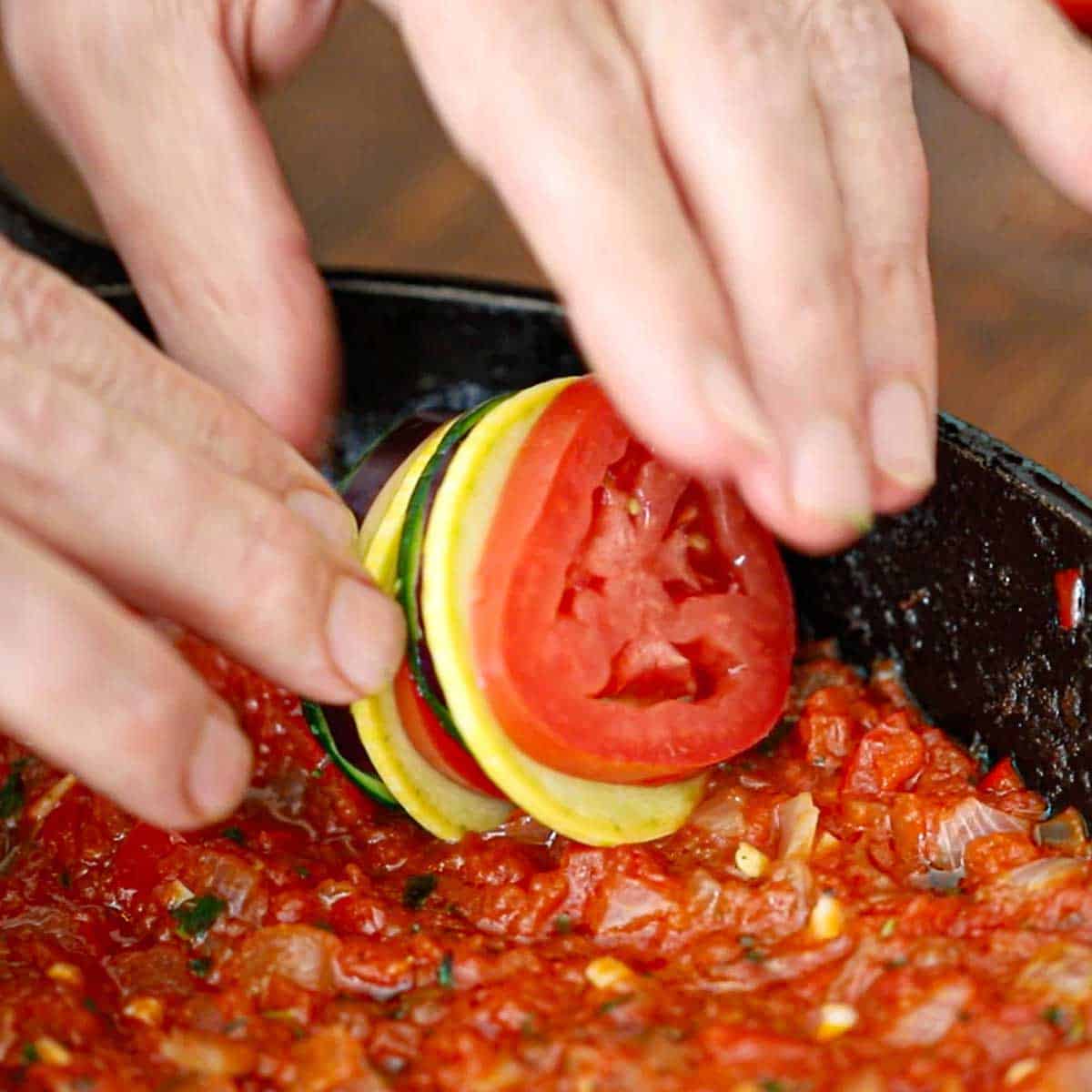 A person placing a stack of vegetables including sliced tomatoes, yellow squash, zucchini, and eggplant on their sides in a cast-iron skillet that has a layer of sautéed tomatoes, onions, and red bell pepper in the bottom of the pan.