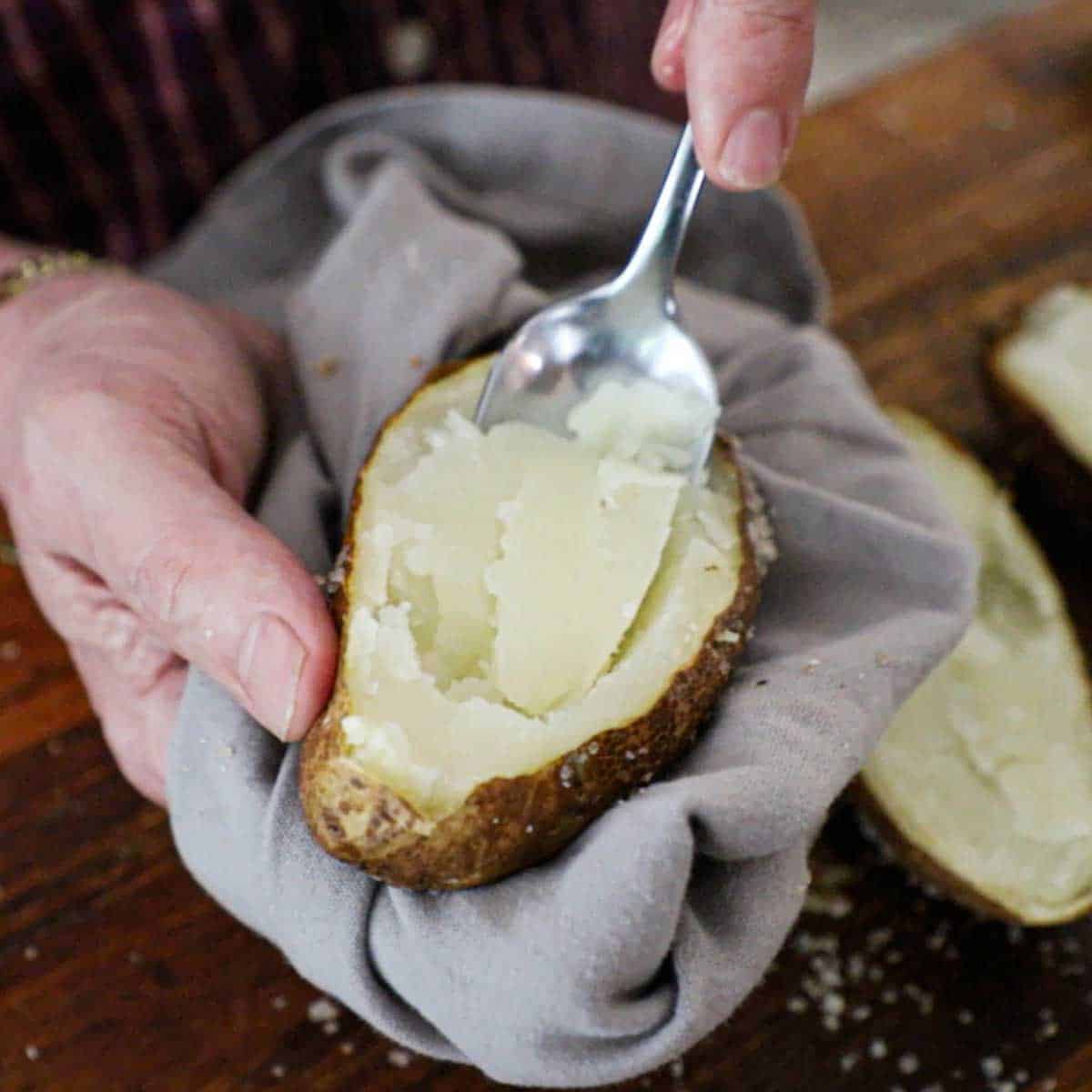 A person using a spoon to remove the inside potato flesh from a baked potato that has been cut in half.