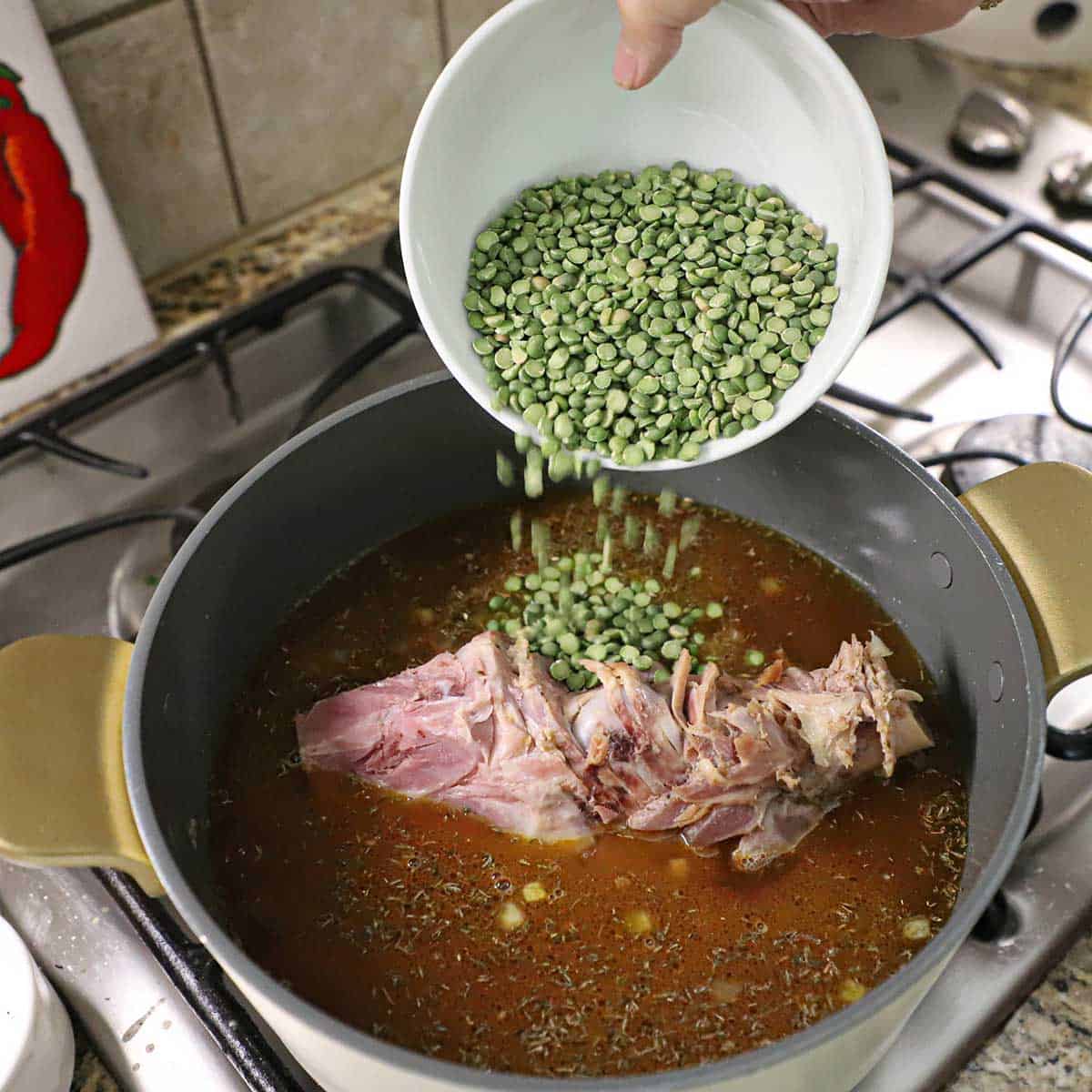 A person transferring split peas from a bowl into a pot filled with a simmering broth and large ham bone on a gas stove.