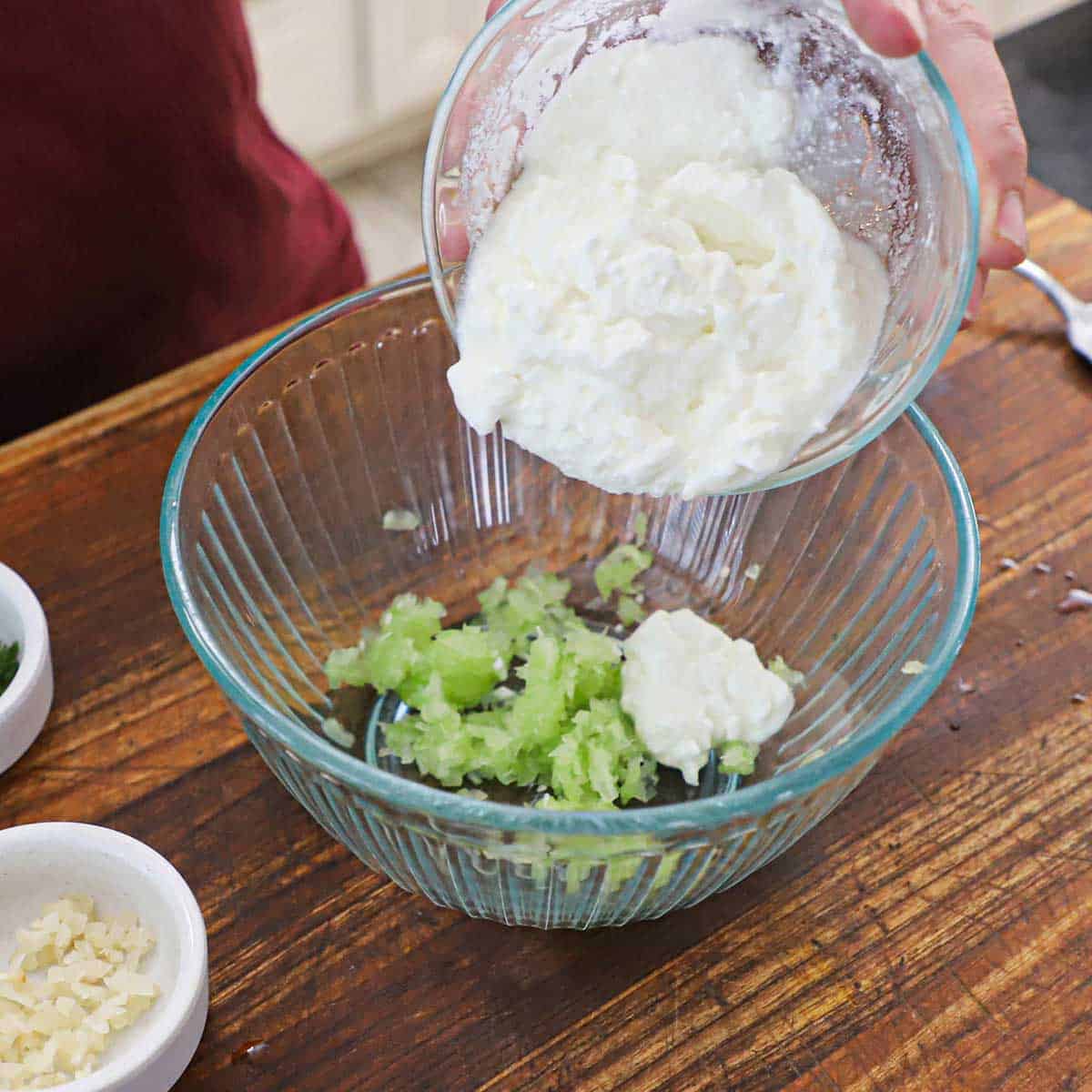 A person transferring plain Greek yogurt from a small glass bowl into another glass bowl that is holding drained, shredded cucumber.