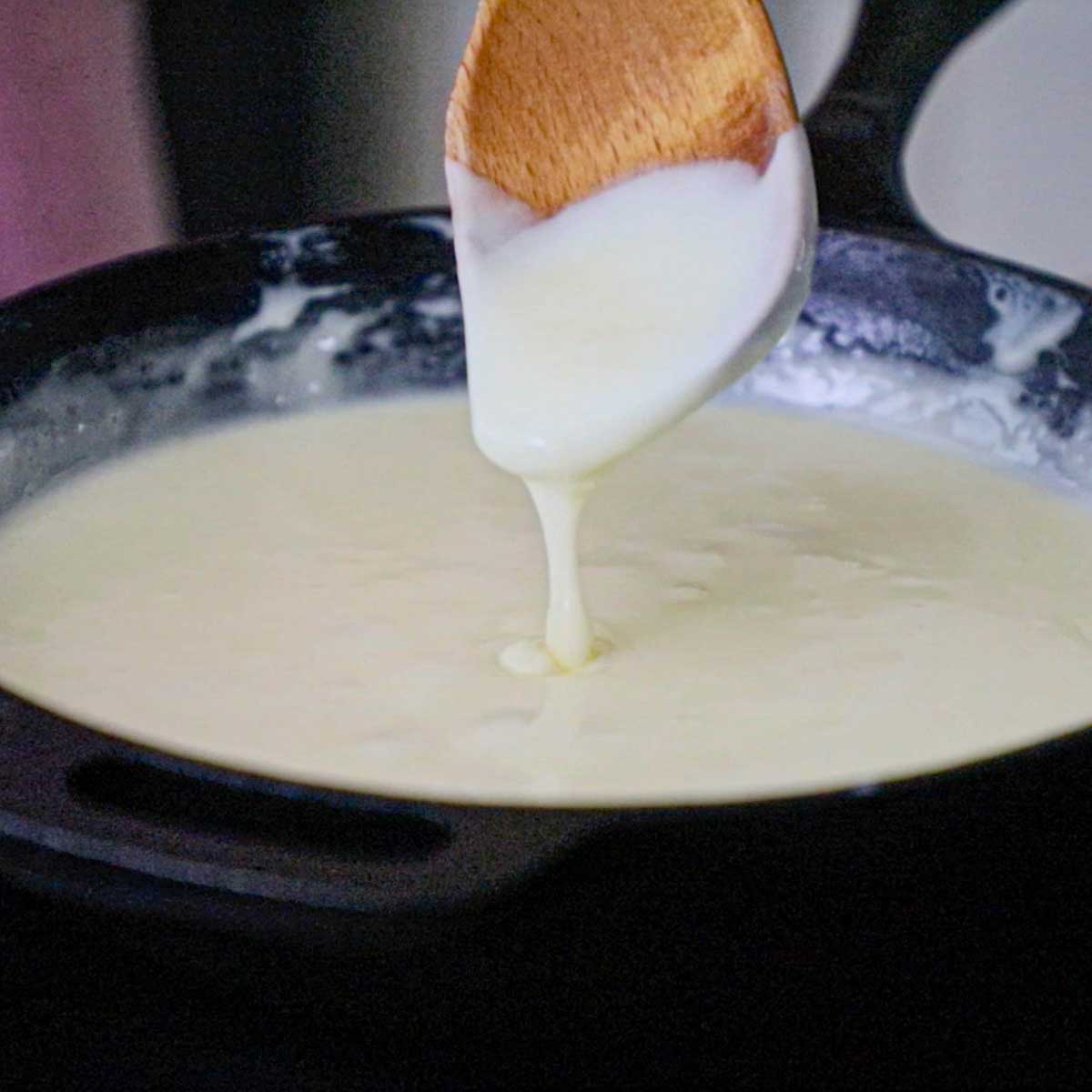 A person raising a wooden spoon form fully melted white American cheese combined with milk and water in a cast-iron skillet.