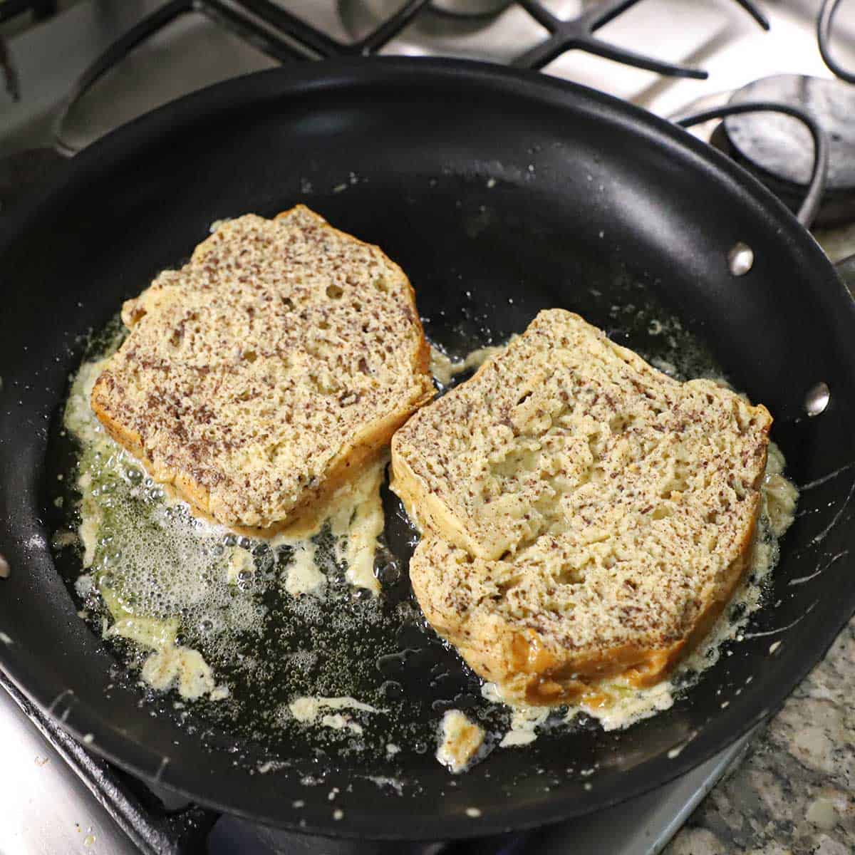 Two custard-soaked slices of brioche being lightly fried in a large black non-stick skillet on a gas stove.