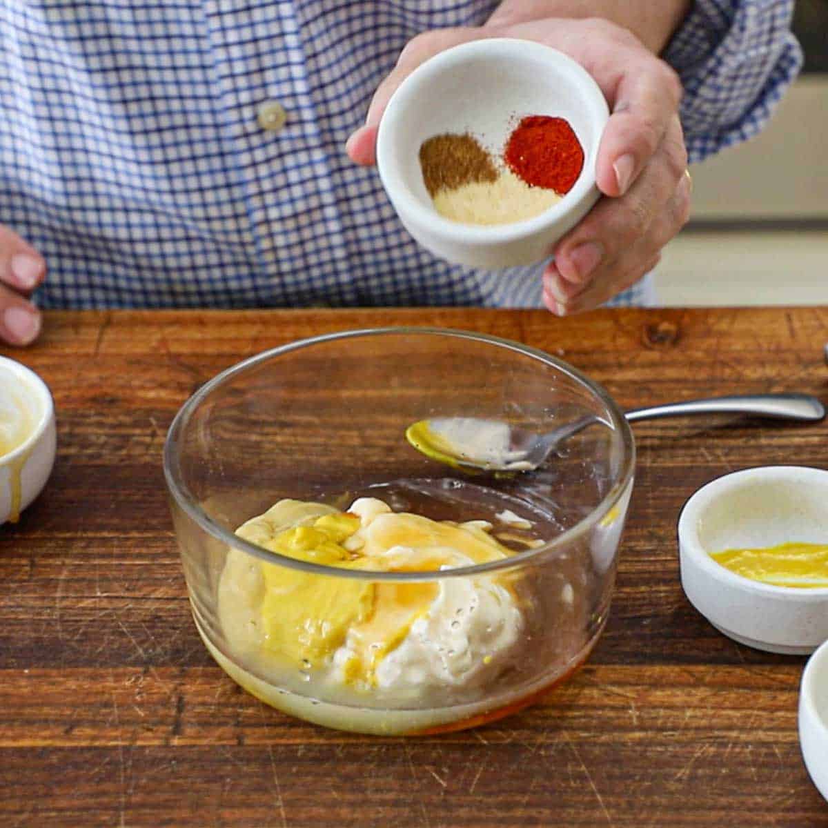 A person holding a small bowl filled with seasonings that he is ready to dump into a bowl filled with mayonnaise, mustard, honey, and lemon juice.