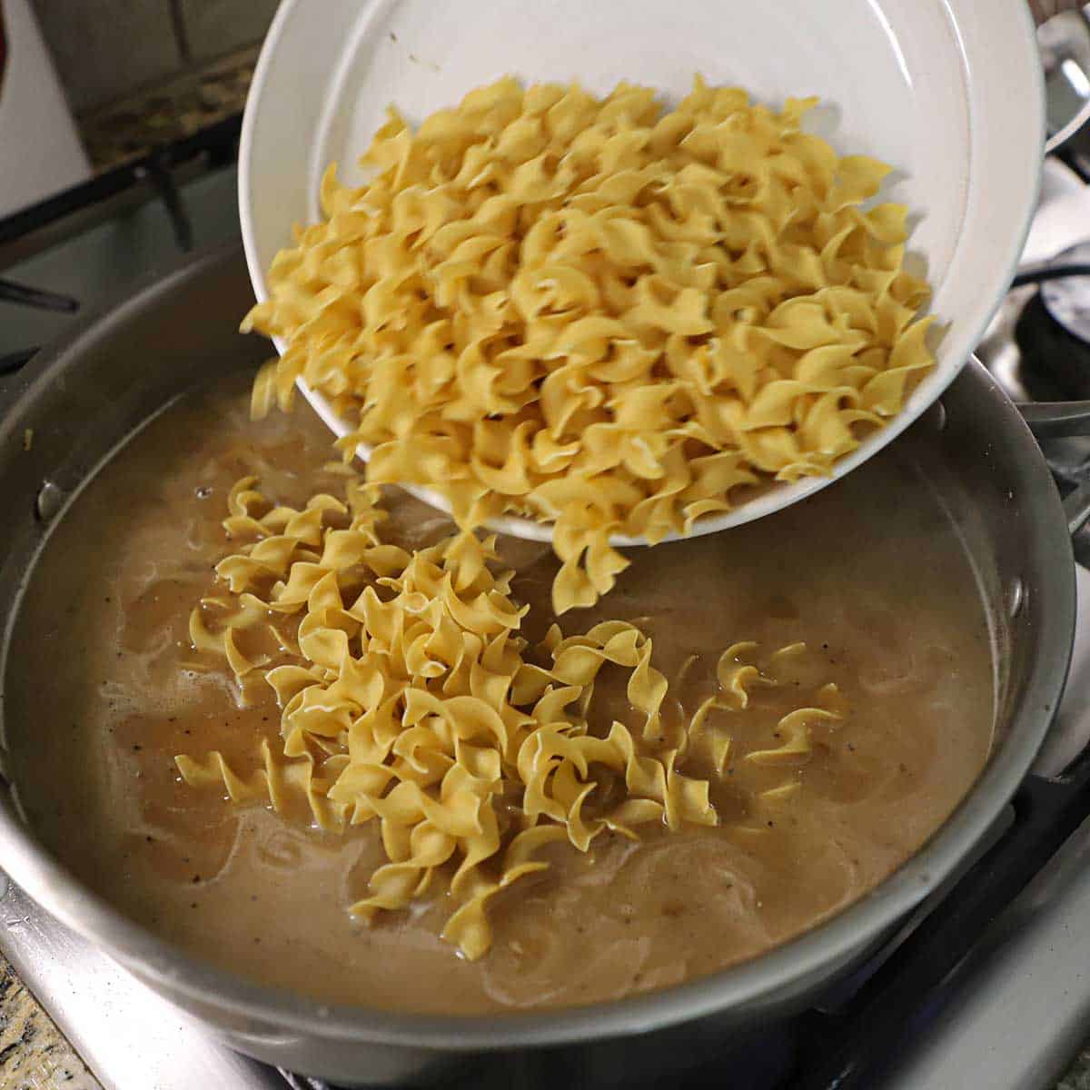 A person dumping uncooked egg noodles into a skillet filled with a simmering brown sauce.