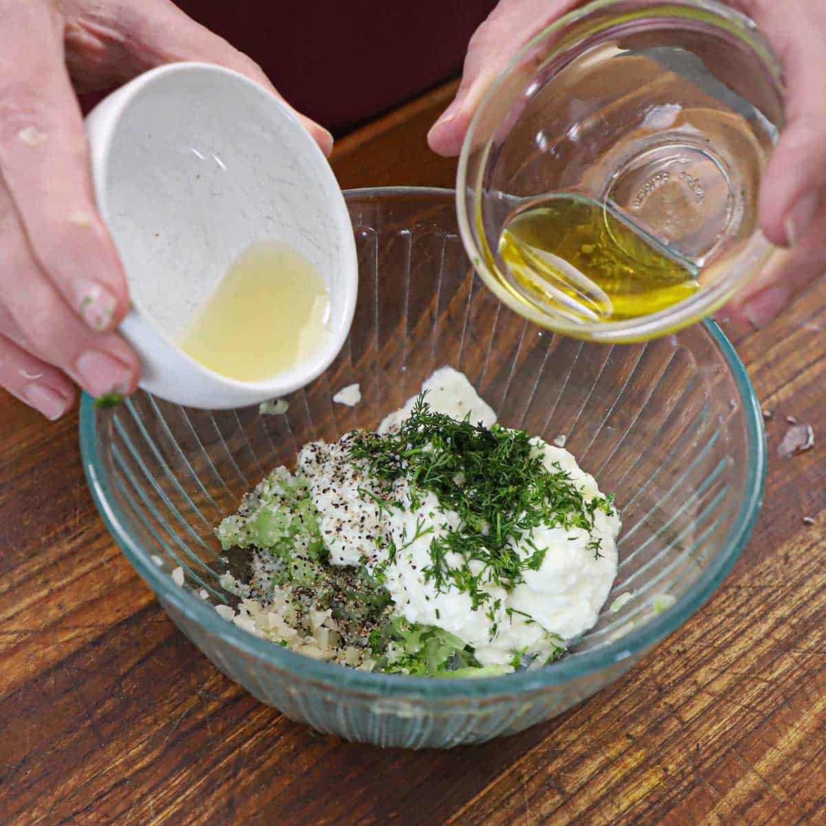 A person holding a small bowl of white wine vinegar in one hand and the other hand holding olive oil both over a glass bowl filled with the ingredients for homemade tzatziki sauce.