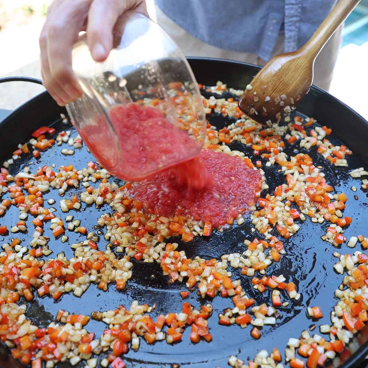 A person transferring grated tomatoes from a glass bowl into a large paella pan that is filled with simmering chopped onions and red bell peppers in hot oil.