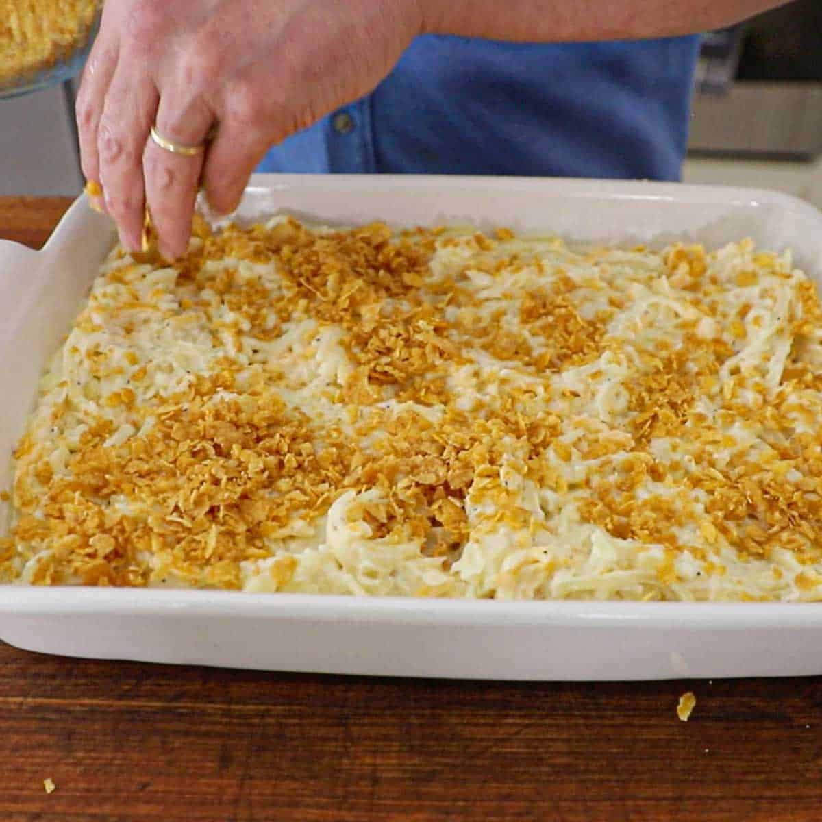 A person sprinkling crushed corn flakes over the top of a white baking dish that is filled with a hashbrown casserole from scratch.