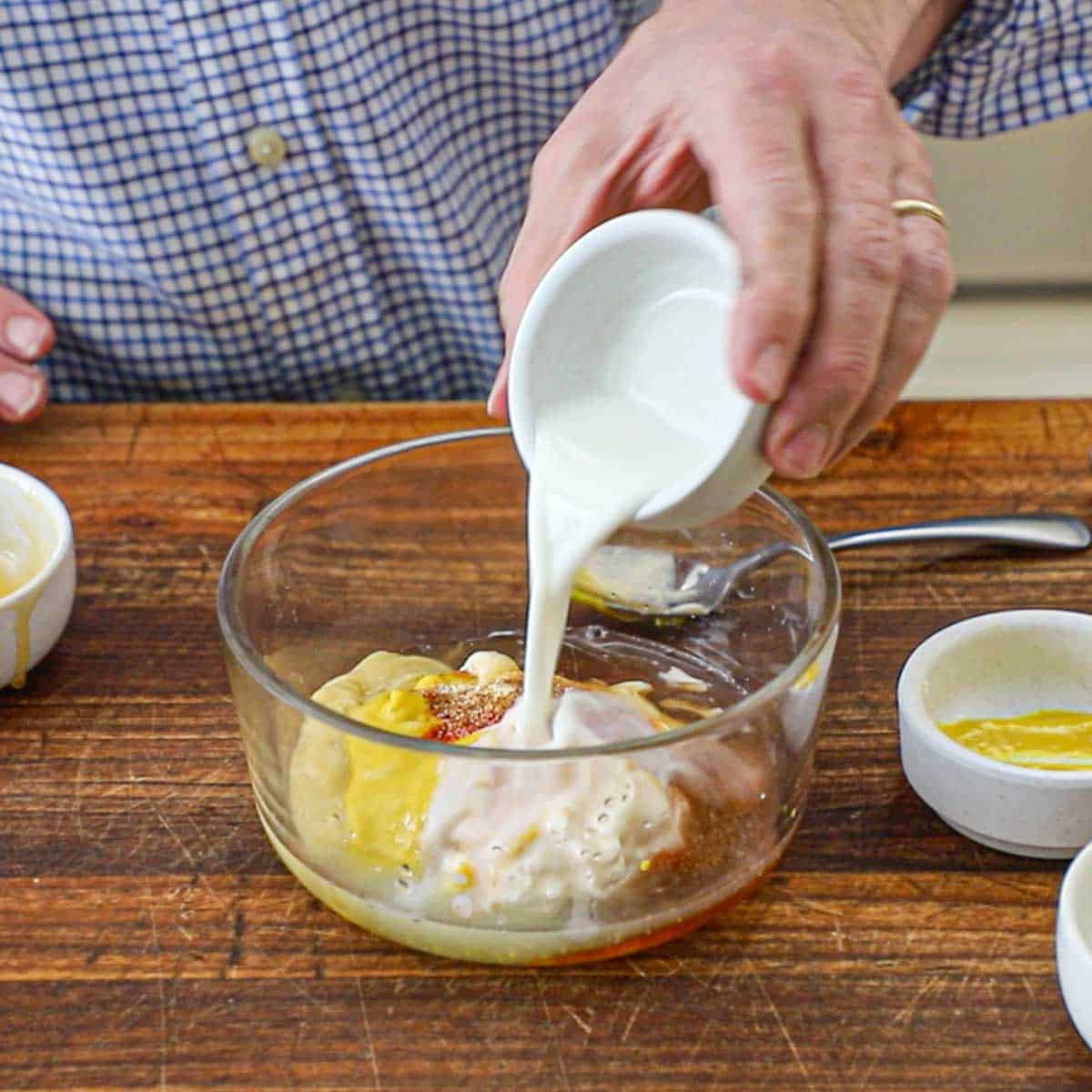 A person pouring a small amount of whole milk from a small bowl into a glass bowl holding the ingredients for Restaurant-Style Honey Mustard Dressing.