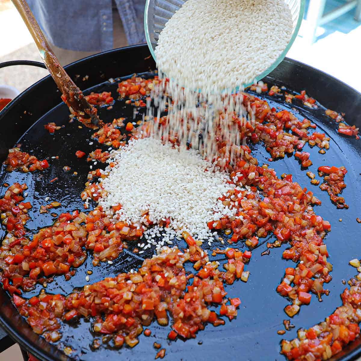 A person transferring short-grain rice from a glass bowl into a large paella pan that is filled with sautéd sofrito.