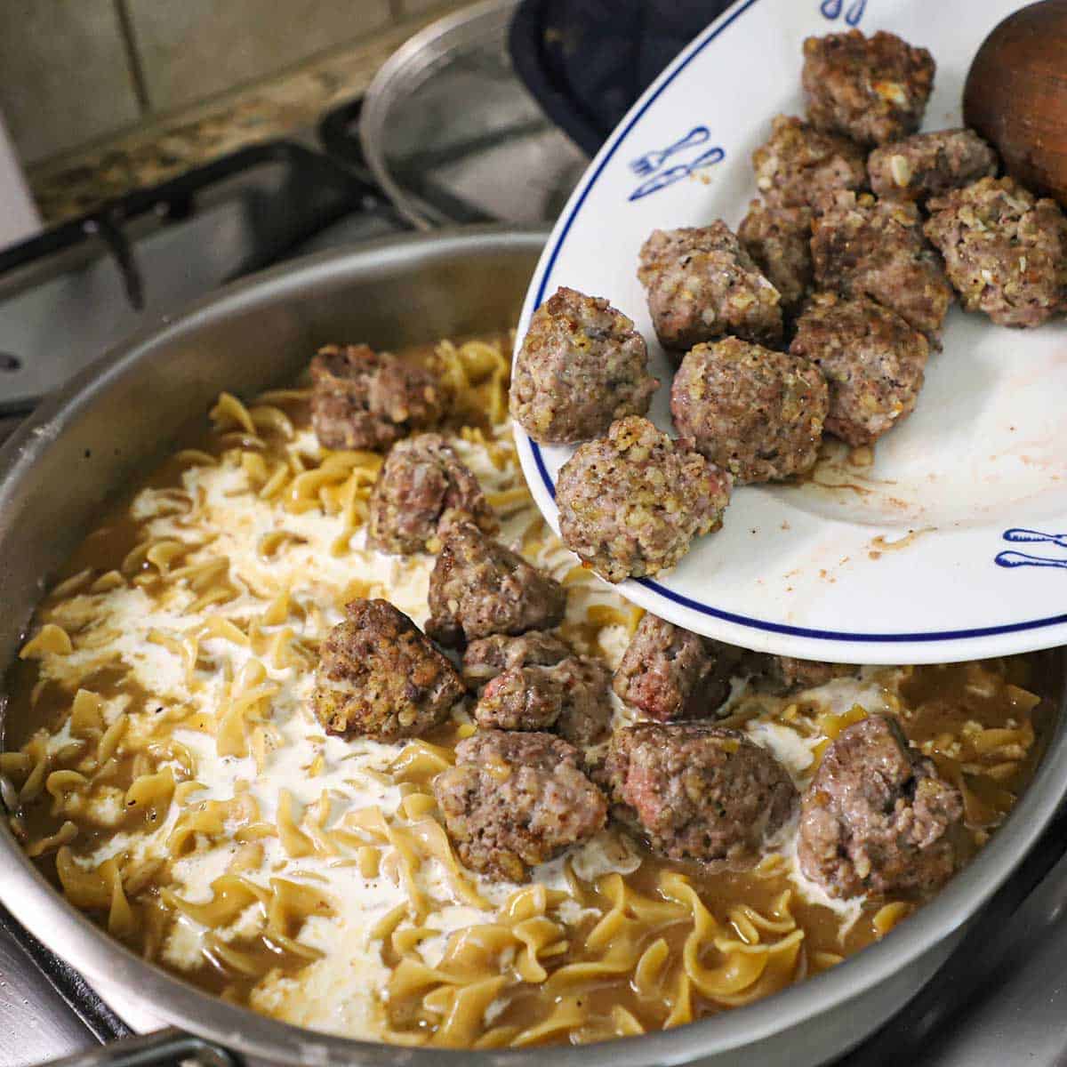 A person transferring partially cooked Swedish meatballs into a large skillet filled with a simmering brown and cream sauce with egg noodles in the sauce.