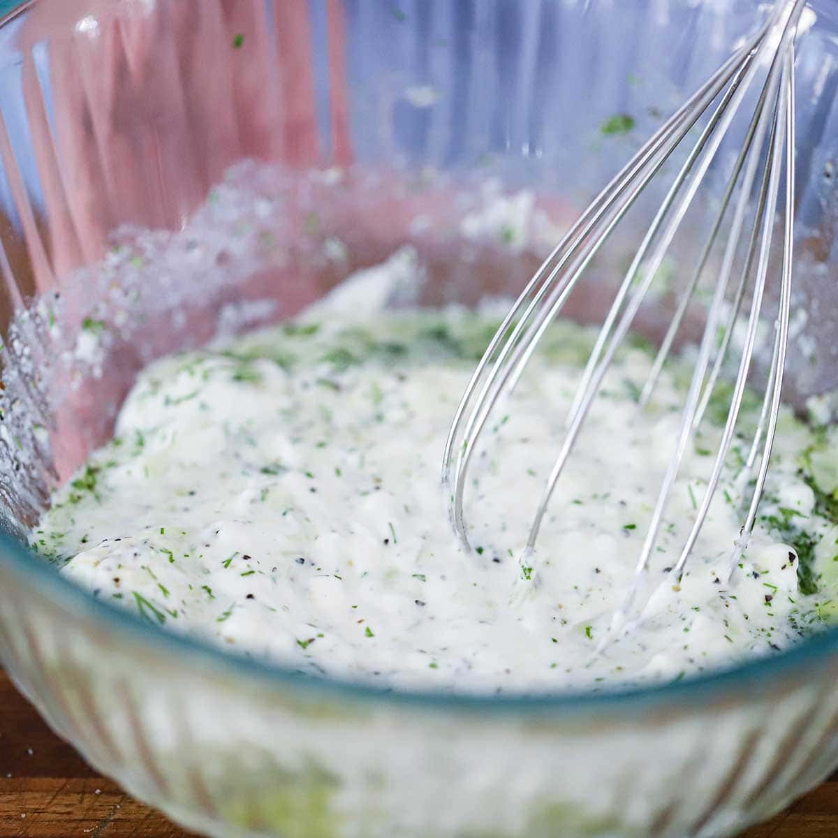 A close-up view of a whisk being used to combine the ingredients for homemade tzatziki sauce in a glass bowl.
