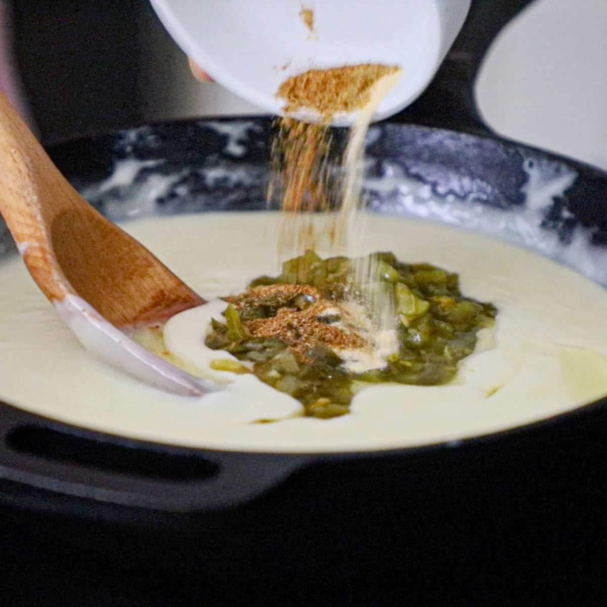 A person dumping dried cumin and garlic powder from a small bowl into a cast-iron skillet that is filled with melty white queso.
