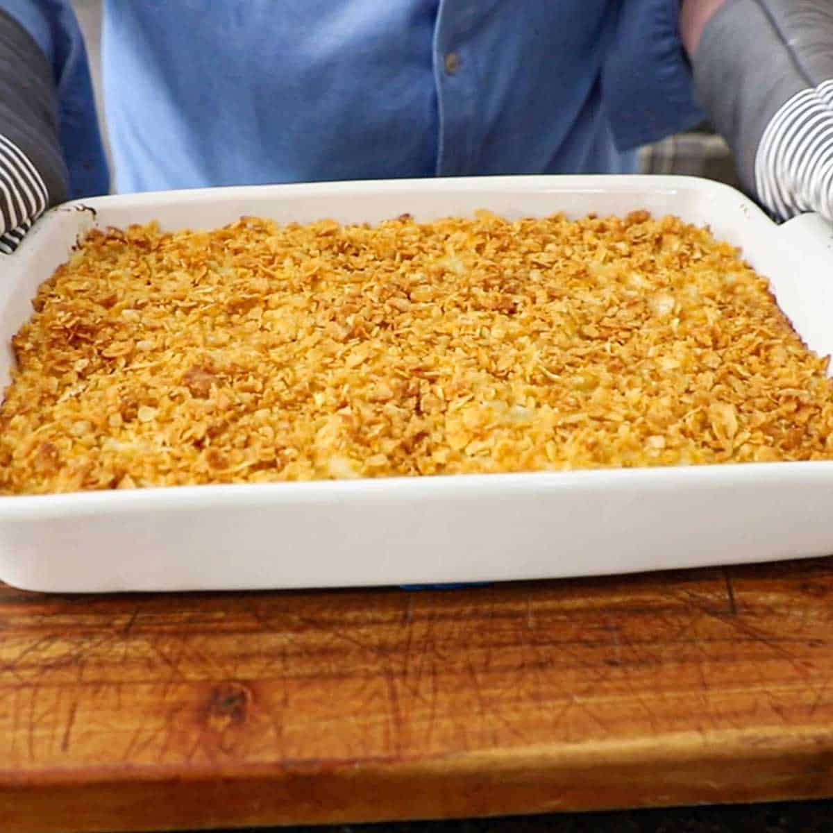 A person with oven mitts holding a white baking dish that contains a freshly baked homemade hashbrown casserole.