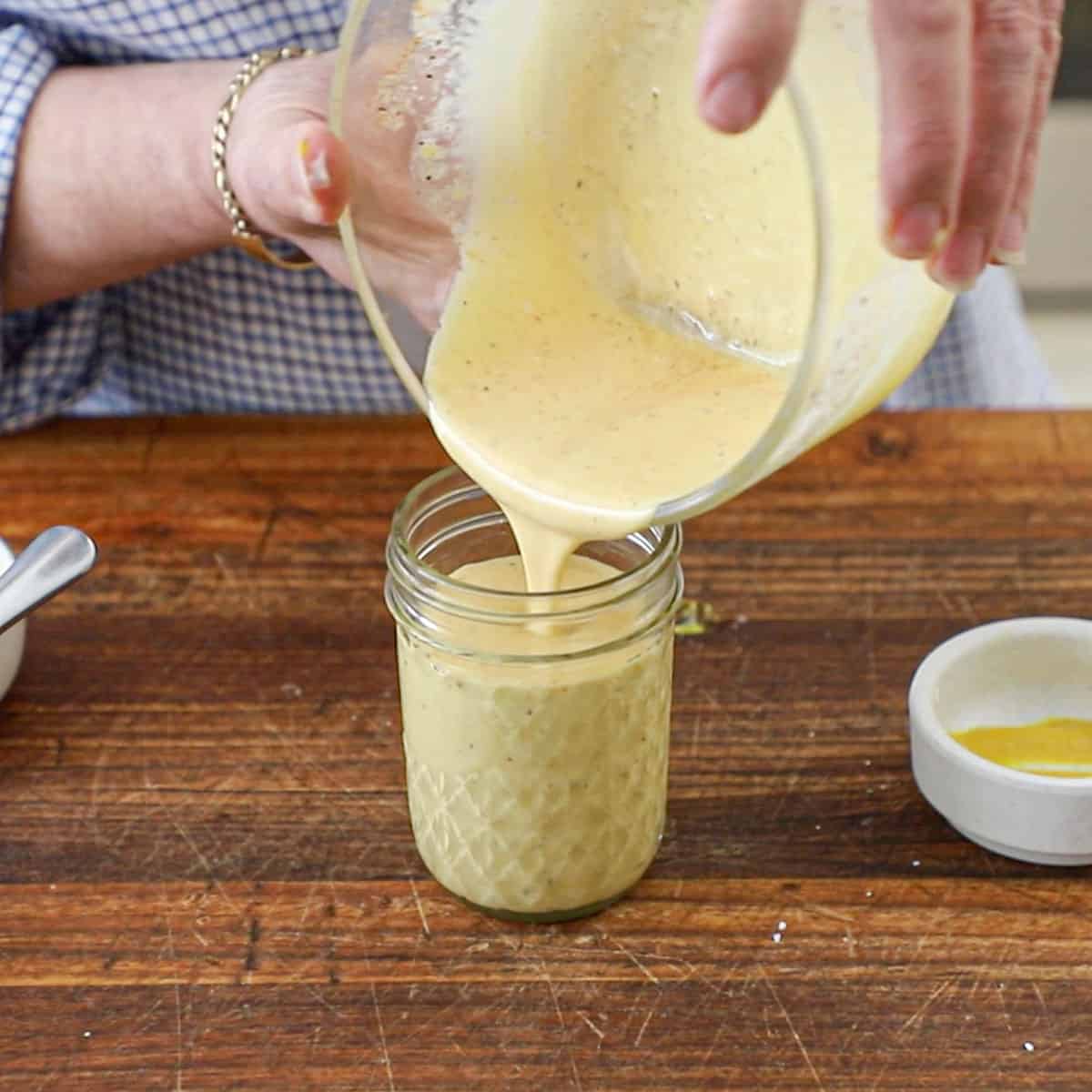 A person pouring freshly prepared Restaurant-Style Honey Mustard Dressing from a glass bowl into a small glass jar on a wooden cutting board.