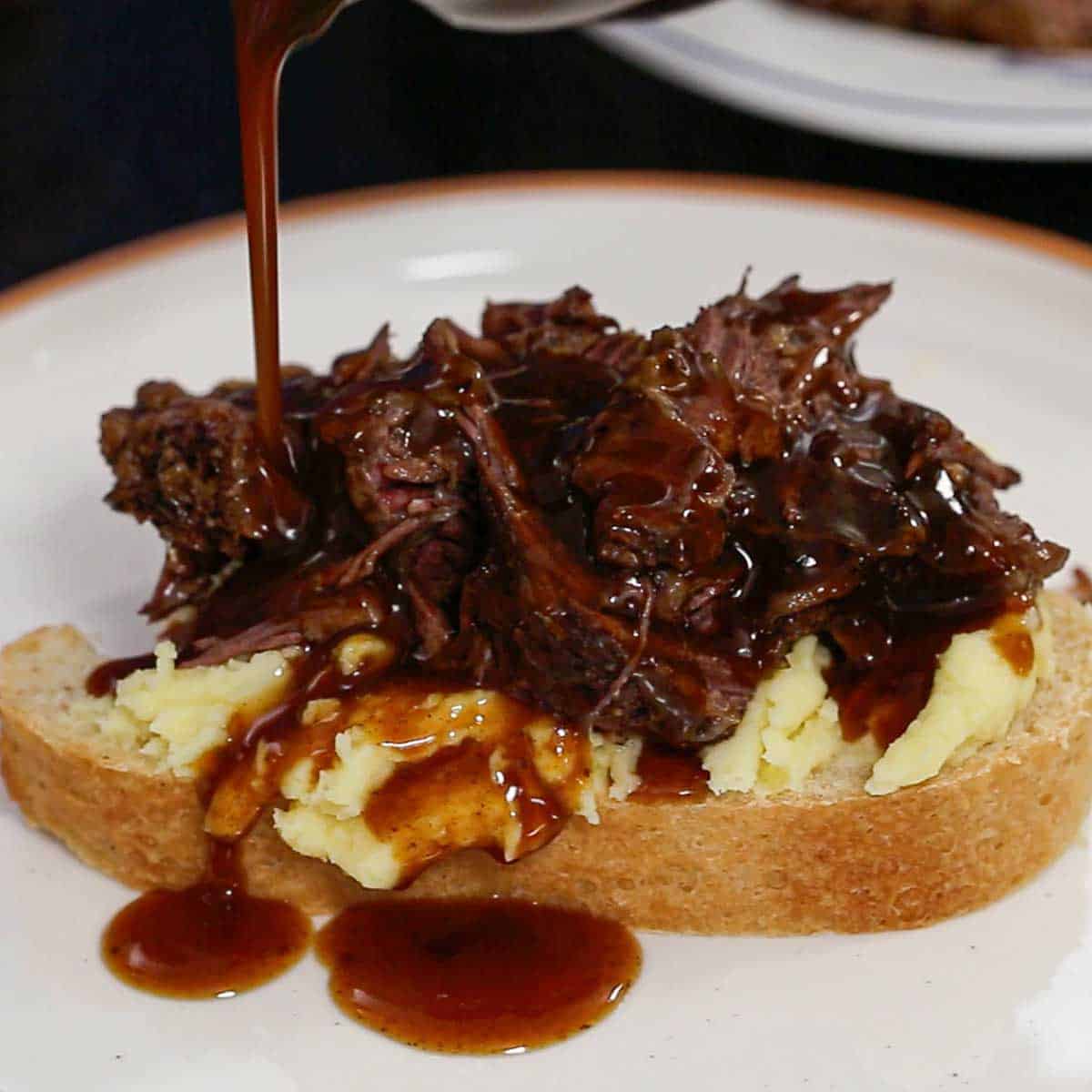 Brown gravy being poured over the top of a serving of slow cooker beef Manhattan including shredded roast beef over mashed potatoes on top of homemade sliced white bread.