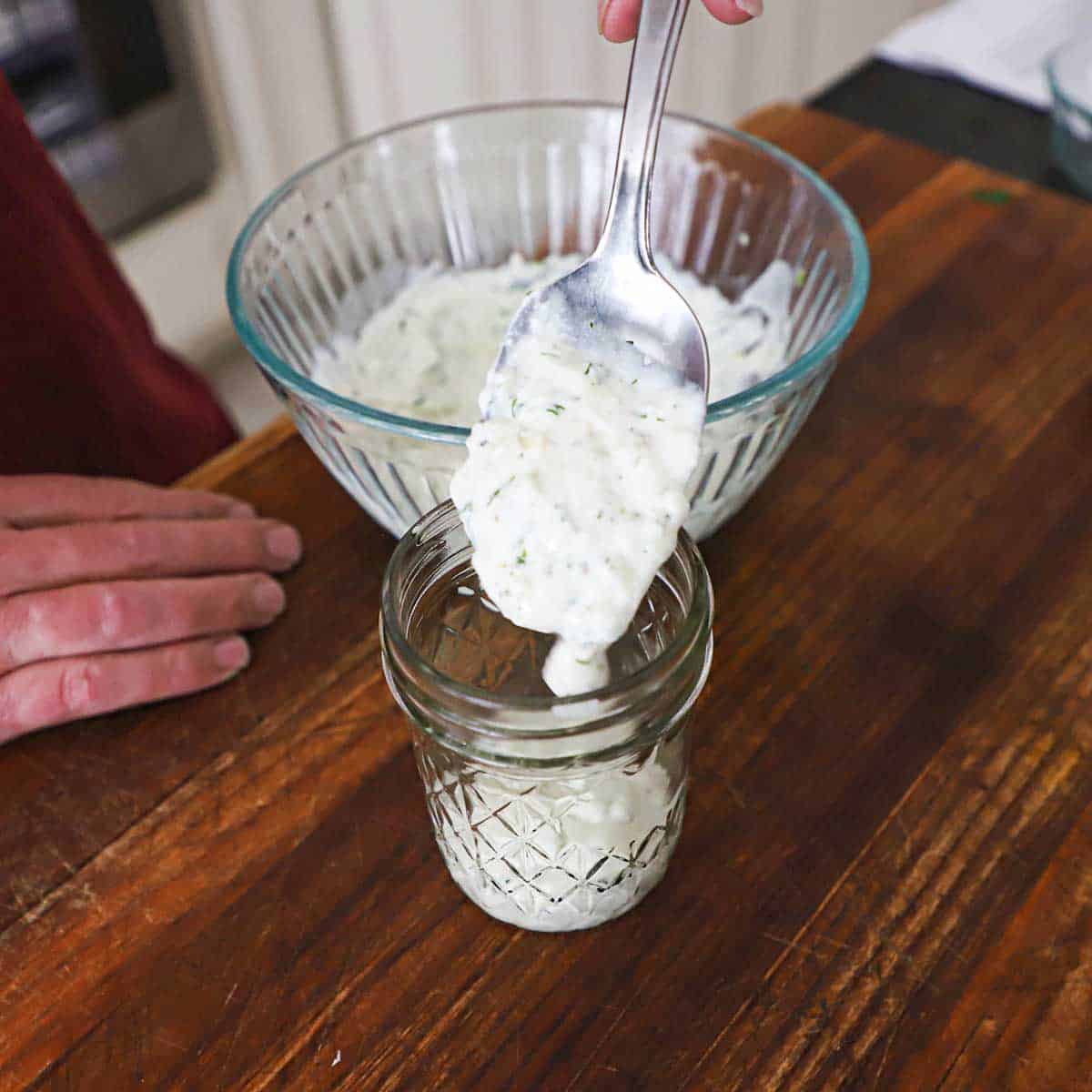 A person using a large spoon to transfer homemade tzatziki sauce from a glass bowl into a small glass jar resting on a wooden cutting board.