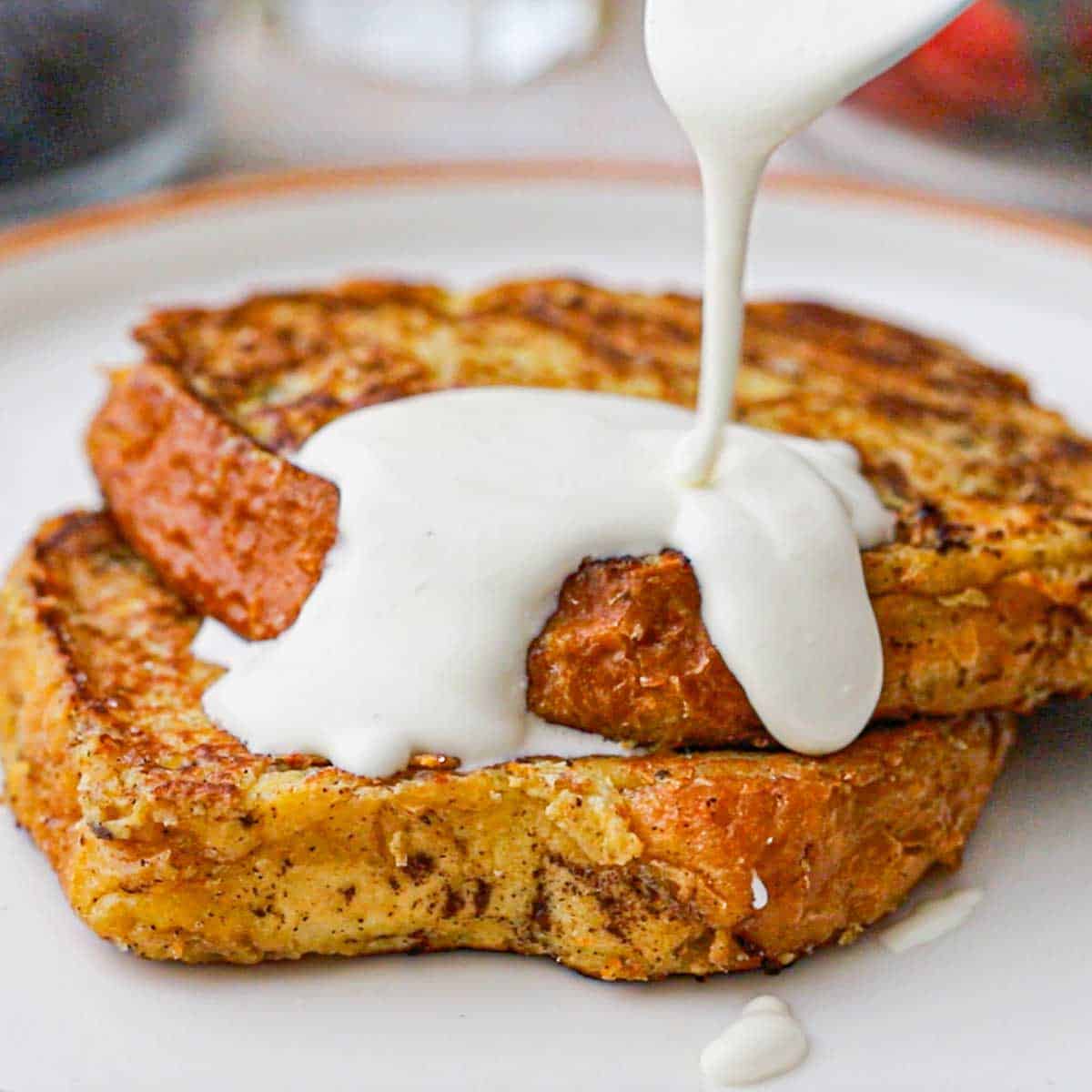 A person using a spoon to drizzle a mascarpone cream sauce over the top of two pieces of caramelized brioche French toast that are stacked on a white dinner plate.