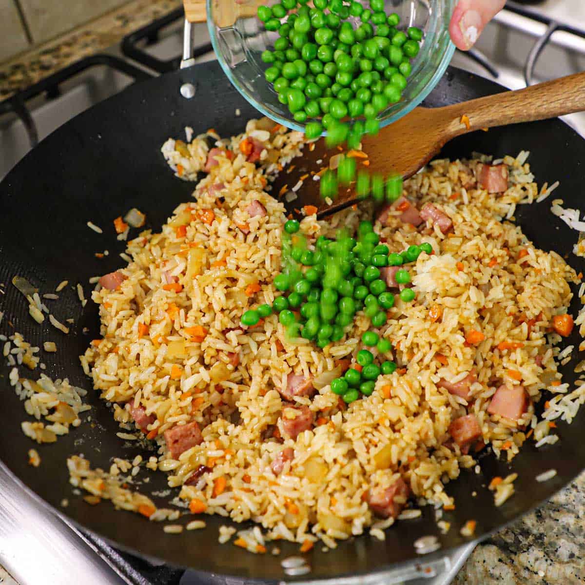 A person transferring frozen sweet peas from a small glass bowl into a large wok filled with simmering leftover ham fried rice.