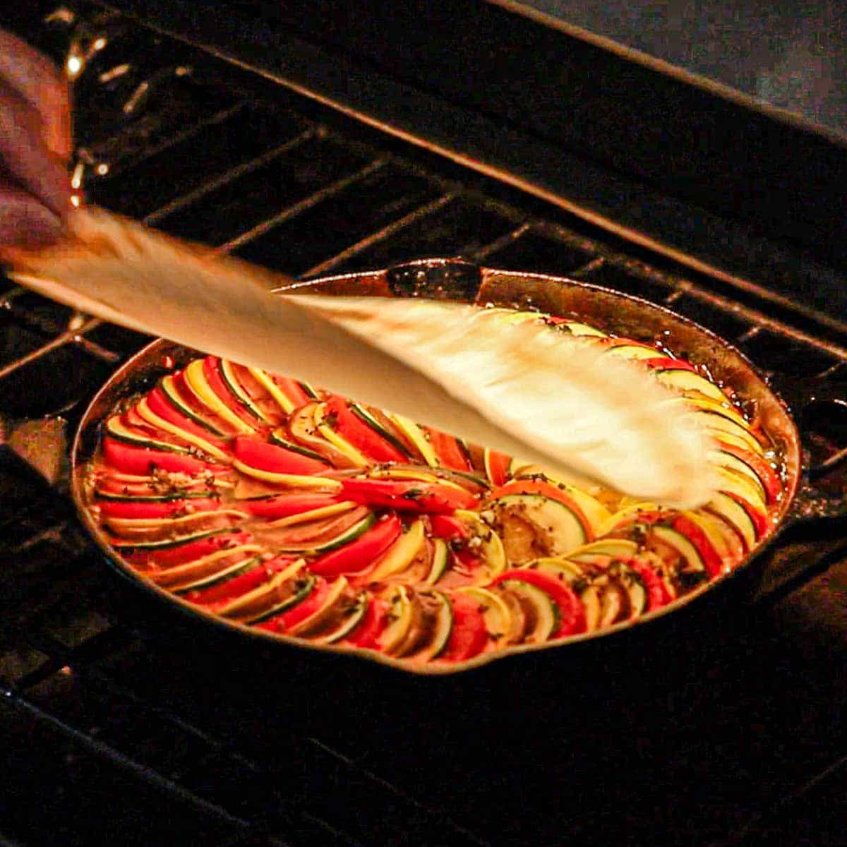 A person removing a circular piece of parchment paper from a cast-iron skillet ratatouille that is being baked in an oven.