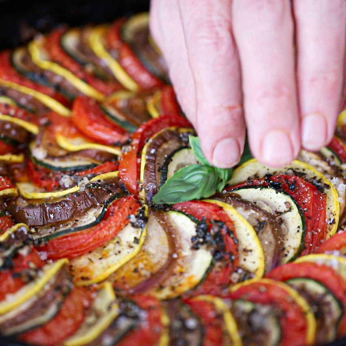 A person placing a sprig of fresh basil leaves in the top center of a freshly baked cast-iron skillet ratatouille.