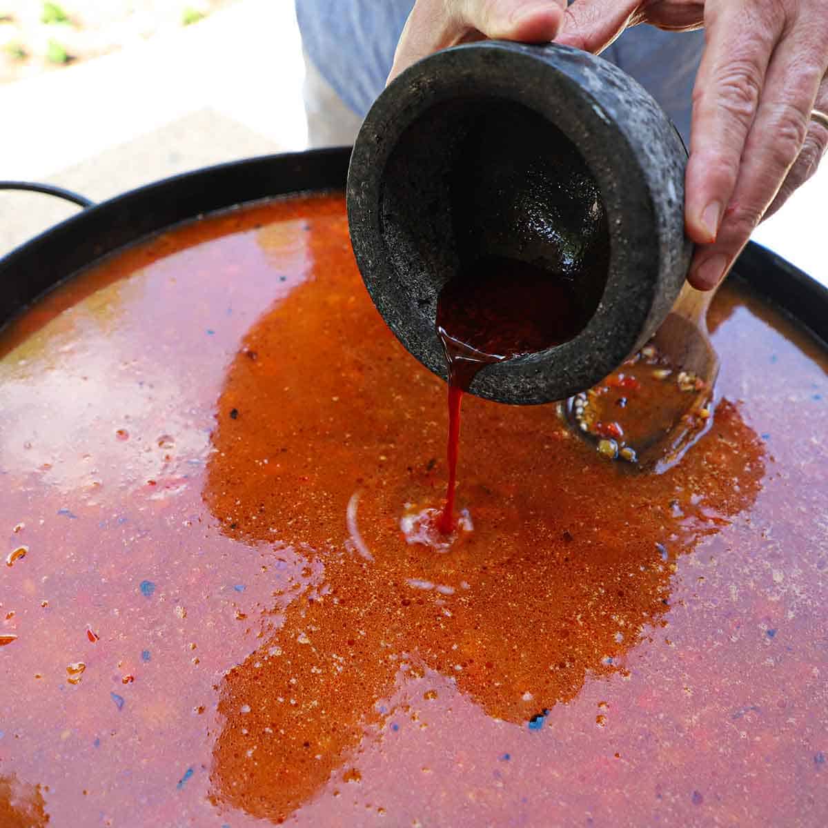 A person pouring saffron-infused shellfish stock from a coral bowl into a paella pan filled with seafood paella that is being simmered.
