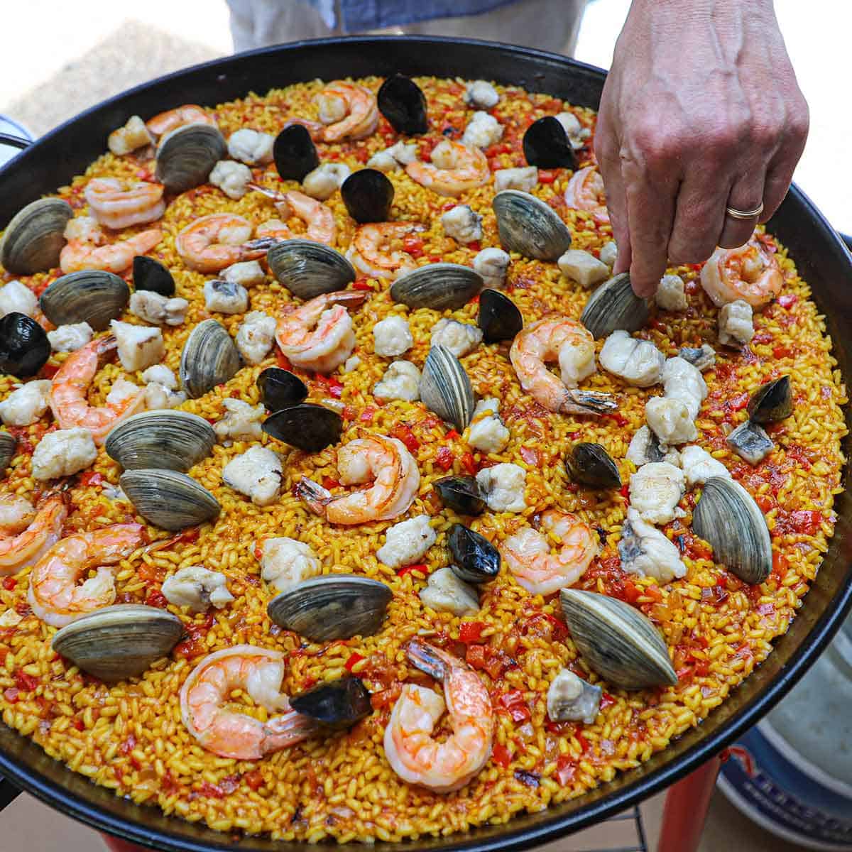A person placing littleneck clams onto the surface of freshly prepared Authentic Spanish Seafood Paella in a large paella pan.