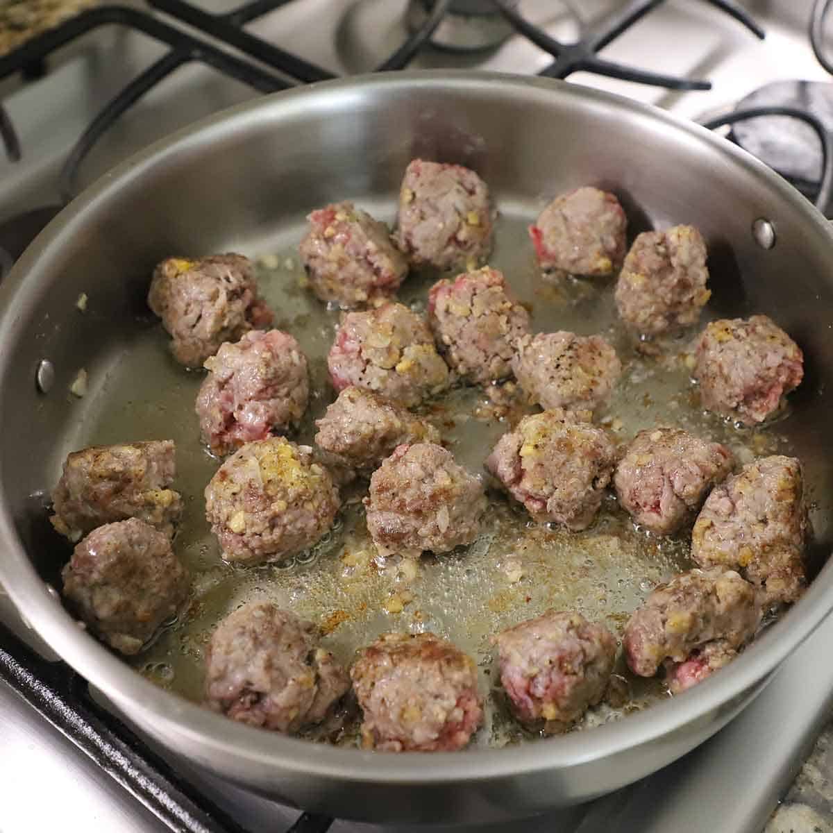 Uncooked Swedish meatballs being seared in a large skillet filled with simmering olive oil on a gas stove.