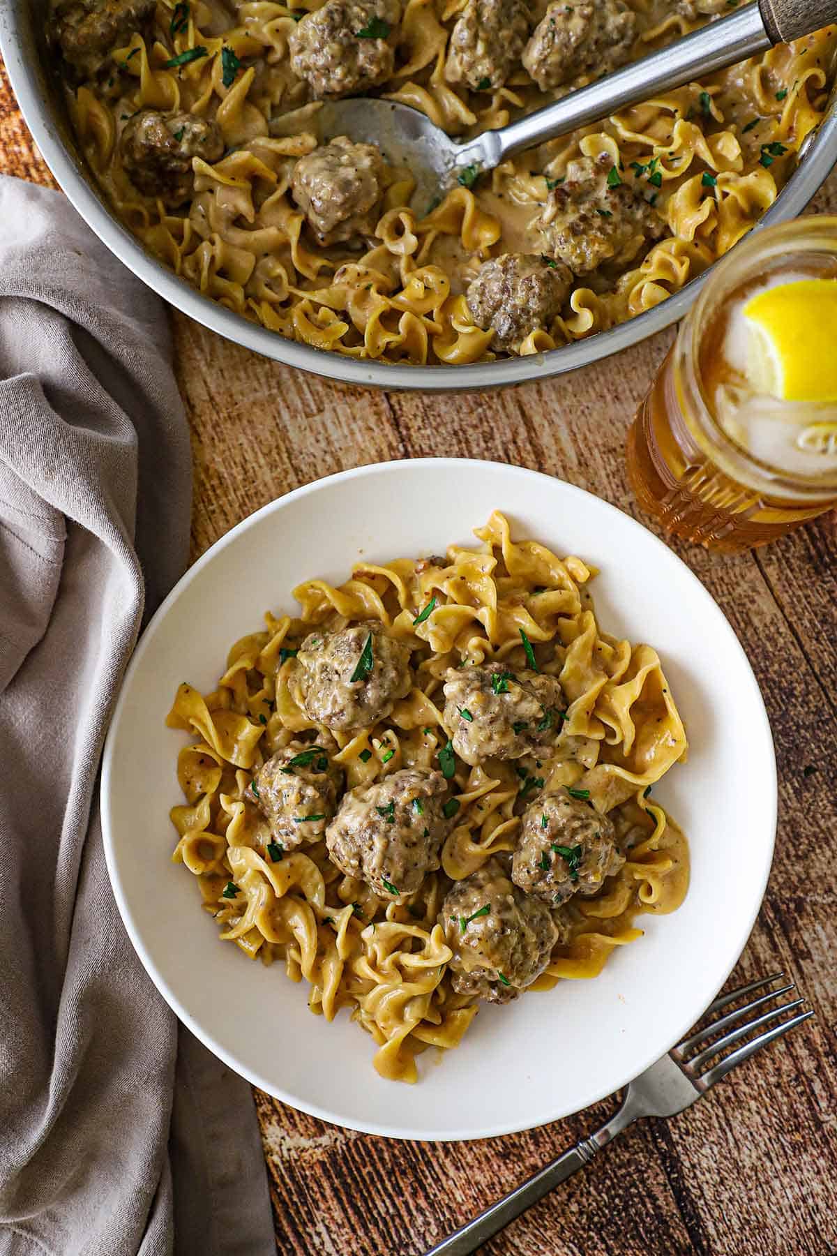 An overhead view of a white pasta bowl filled with a serving of Swedish meatball pasta sitting next to a large skillet filled with the same.