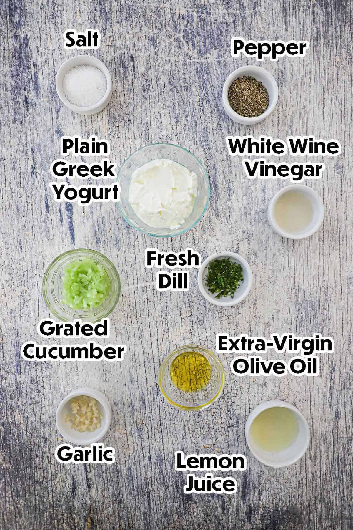 An arrangement of ingredients for homemade tzatziki sauce on a grey wooden background including Greek yogurt, grated cucumber, vinegar, fresh dill, garlic, lemon juice, oil, and seasonings.