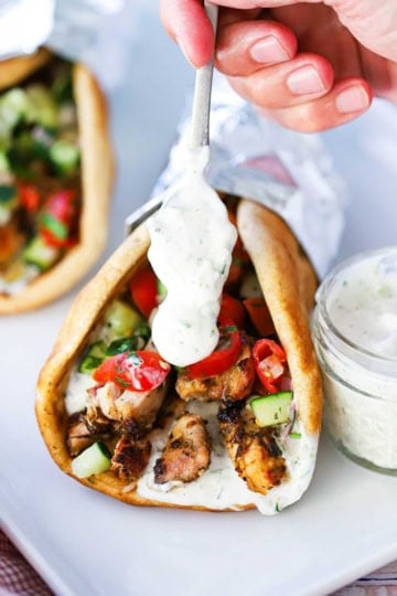 A person using a small spoon to drizzle homemade tzatziki over the end of a wrapped chicken gyro.